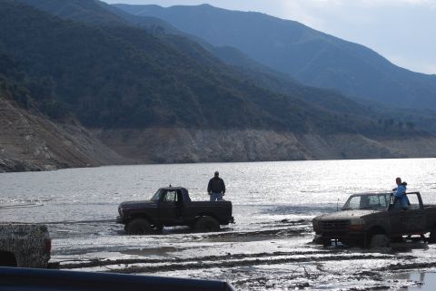1996 Ford Bronco - Two trucks are stuck in the mud near a body of water with mountains in the distance.