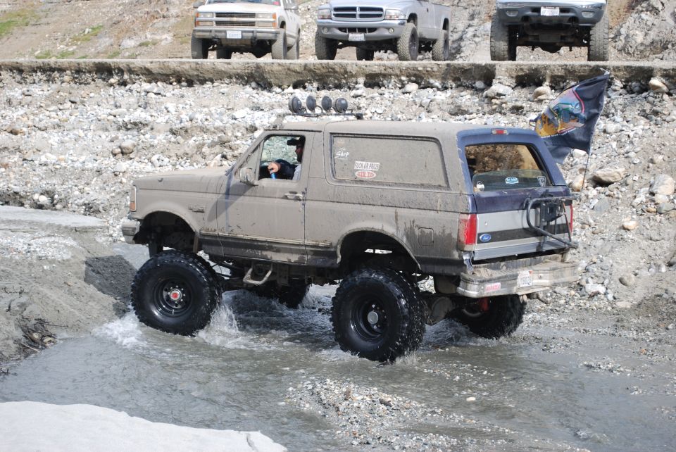 Muddy tan and blue Ford Bronco with lifted suspension and large tires driving through water