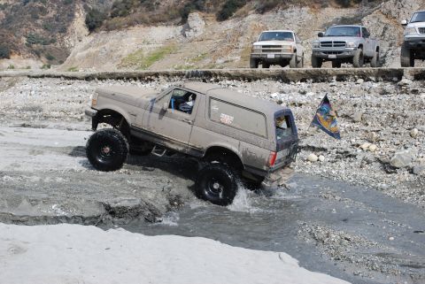1996 Ford Bronco - A lifted 1996 Ford Bronco navigates a rocky stream bed with other trucks visible on the bank.