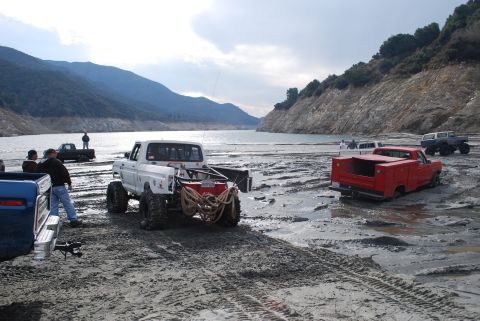 1996 Ford Bronco - Multiple trucks navigate a muddy, lakeside area with steep hills under a cloudy sky.