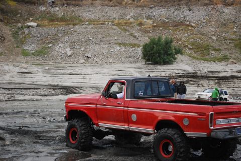 1996 Ford Bronco - A lifted red pickup truck with a black top navigates a muddy, off-road terrain, with other vehicles visible in the distance.