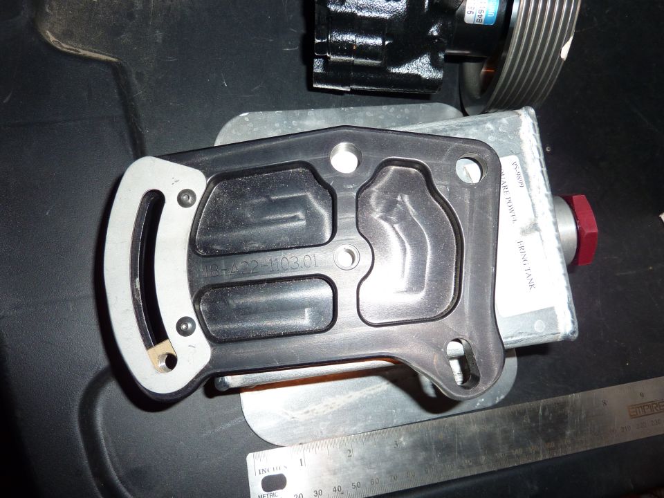 Black gasket with metal trim and stamped numbers sits on a metal plate next to a power steering pump