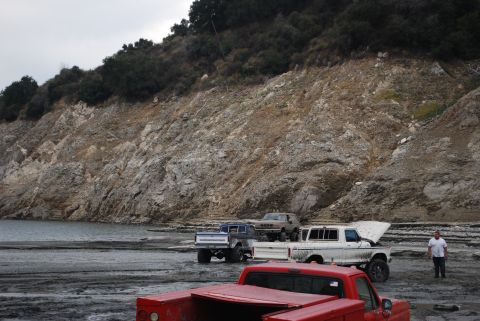 1996 Ford Bronco - Several trucks and SUVs are parked on a muddy shore next to a rocky hillside and a body of water.