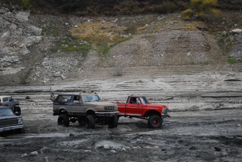 1996 Ford Bronco - Two , lifted trucks navigate a muddy, off-road landscape under a barren hillside.