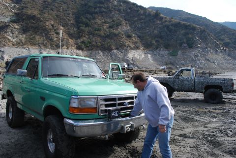 1996 Ford Bronco - A man examines a green Ford Bronco, with another muddy truck in the background on a dirt terrain.