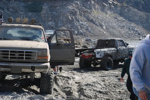 1996 Ford Bronco - Two heavily mud-covered trucks are parked on a dirt and rocky area near a body of water.