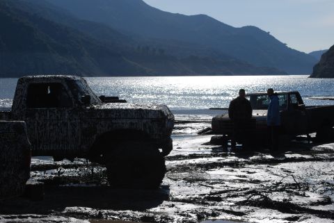 1996 Ford Bronco - Muddy trucks sit by the water's edge with mountains behind them.