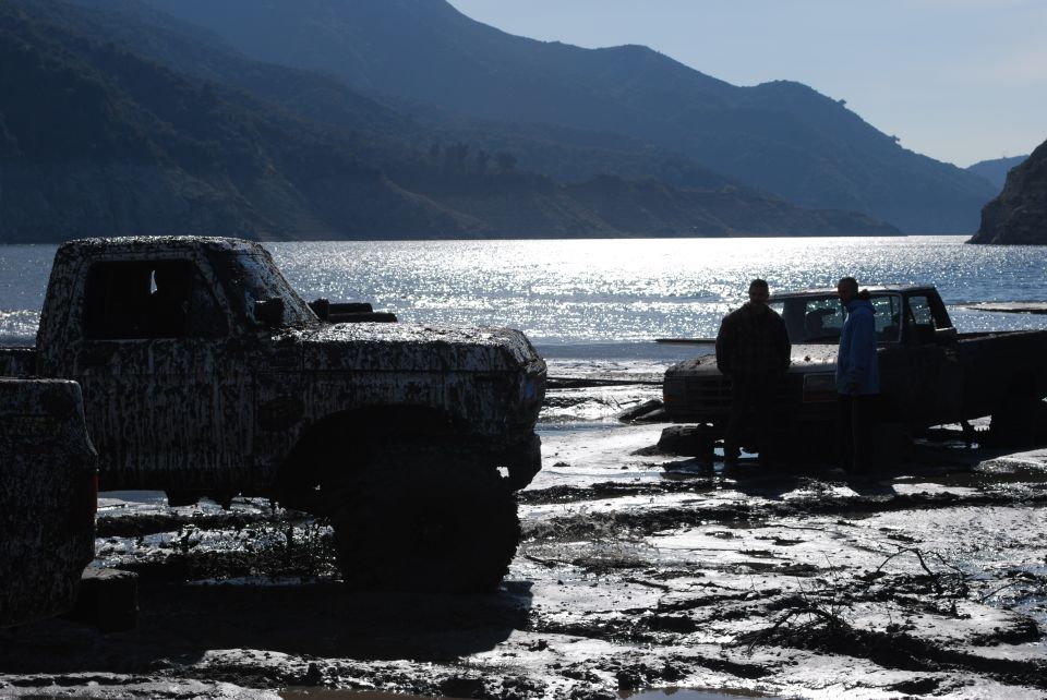 Two mud-covered trucks parked on a muddy shore next to a body of water with mountains in the background
