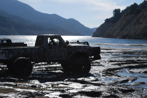 1996 Ford Bronco - Two mud-caked pickup trucks are stuck in a muddy area next to a large body of water with mountains in the distance.