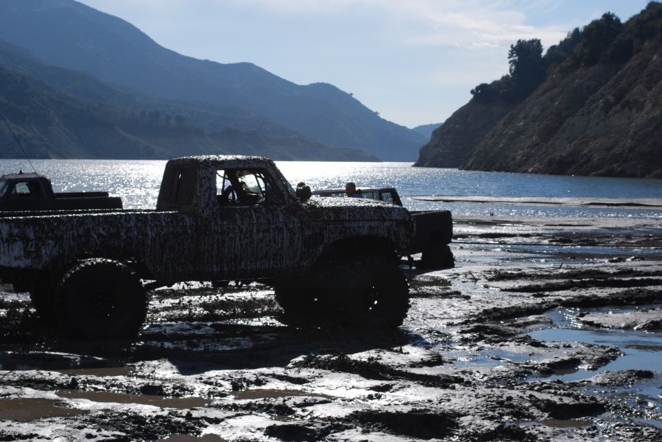 Mud-covered pickup truck stuck in mud near a body of water, with mountains in the background