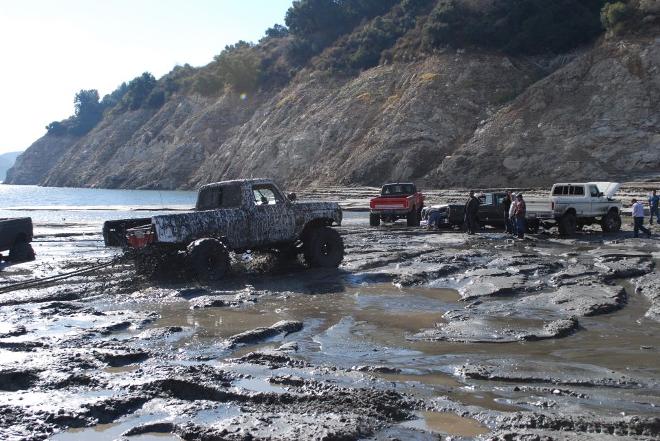 Several trucks stuck in mud near a body of water with a rocky hillside in the background