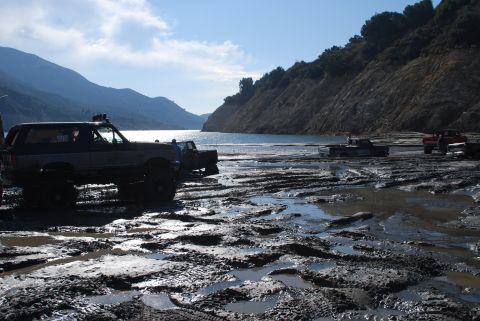 1996 Ford Bronco - Several off-road vehicles navigate a muddy terrain beside a large lake under a clear sky.
