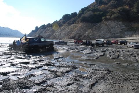 1996 Ford Bronco - Off-road vehicles driving through mud near a lake with a mountainous backdrop.