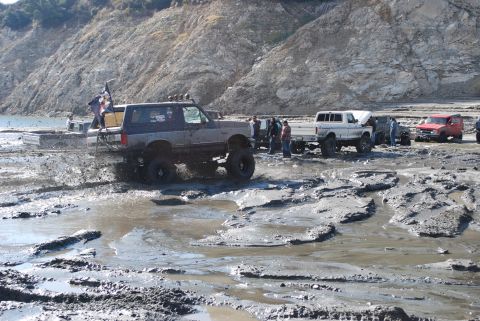 1996 Ford Bronco - Several off-road vehicles navigate a muddy terrain with a rocky hillside in the background.