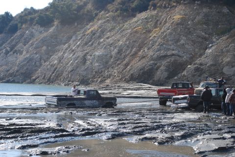 1996 Ford Bronco - Multiple trucks are stuck in deep mud, with one being towed by another near a lake and a steep embankment.