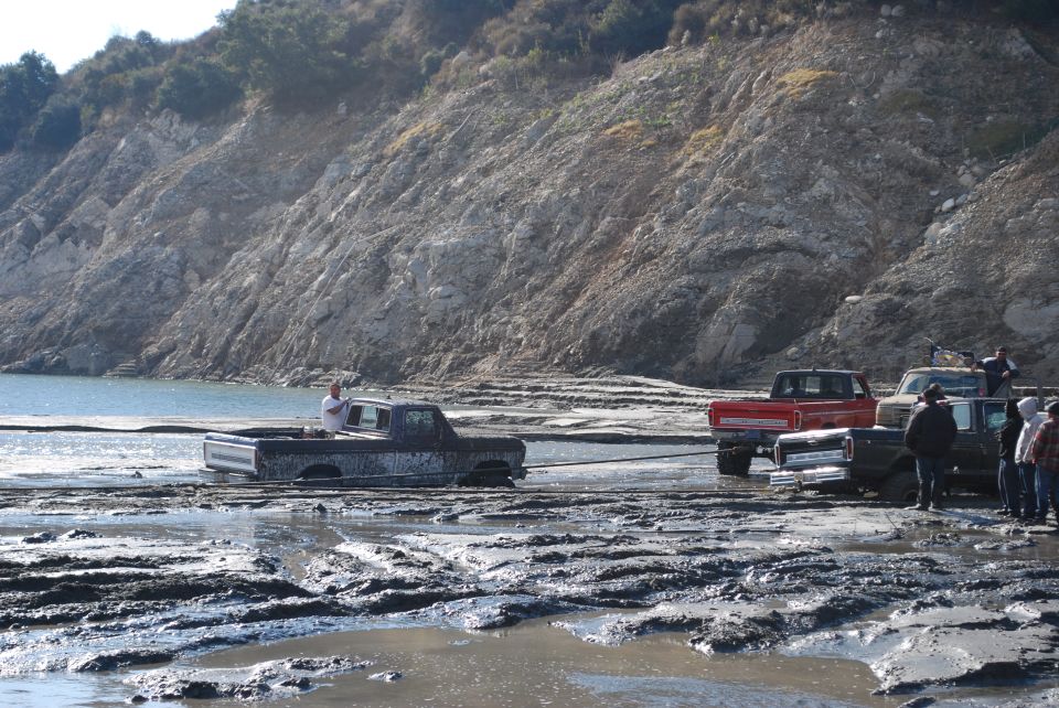 Muddy scene with several trucks stuck in thick mud near a body of water and a rocky hill