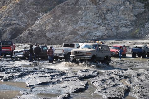 1996 Ford Bronco - Several off-road vehicles navigate a muddy terrain, with a gray Ford Bronco prominently splashing through the water.