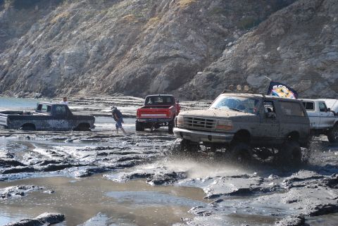 1996 Ford Bronco - Several off-road vehicles navigate a muddy terrain near a rocky hillside.