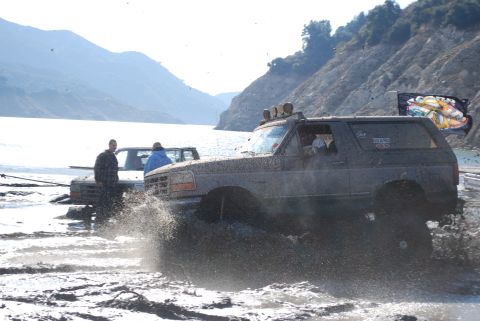 1996 Ford Bronco - This dark SUV is navigating through a muddy terrain, kicking up a large splash as it goes. Another vehicle is visible behind it.