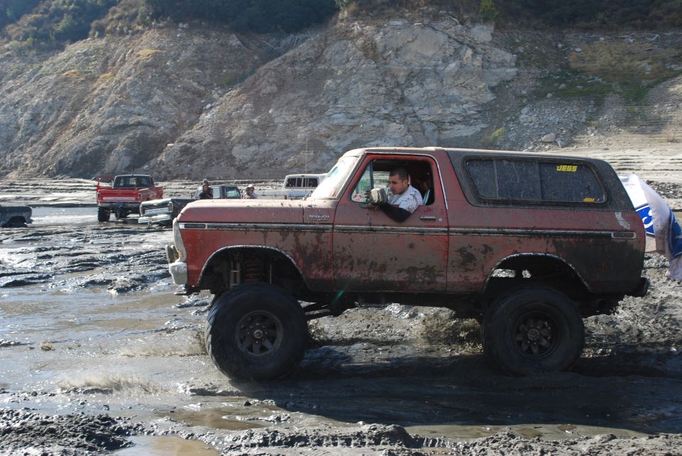 Mud-covered 1978 Ford Bronco with lifted suspension driving through a muddy terrain. Another red truck is visible in the background