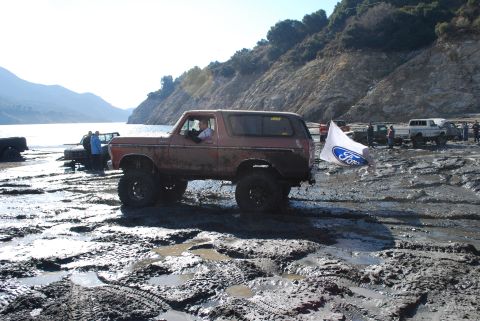 1996 Ford Bronco - A lifted, older model Ford Bronco navigates a muddy terrain near a body of water, with other vehicles and people in the background.