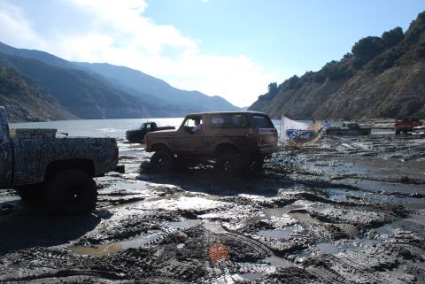 1996 Ford Bronco - Multiple off-road vehicles navigate a muddy terrain near a body of water under a clear sky.