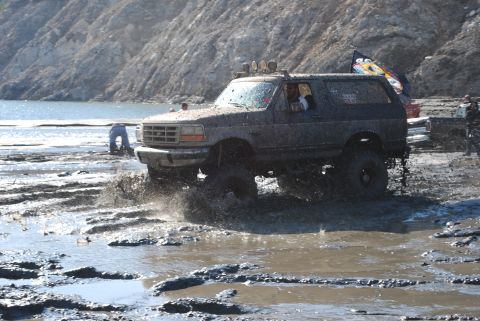 1996 Ford Bronco - This lifted SUV navigates a muddy terrain, kicking up water and mud as it goes. The 'SUPERMOTORS' logo is visible at the bottom right.
