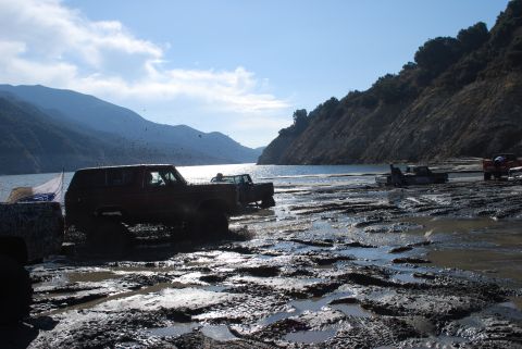 1996 Ford Bronco - Several vehicles are navigating a challenging muddy terrain next to a large body of water under a partly cloudy sky.