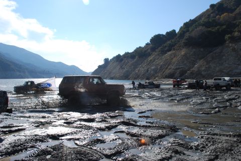 1996 Ford Bronco - Several vehicles navigate a muddy terrain near a body of water under a clear sky.