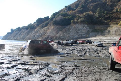 1996 Ford Bronco - Multiple off-road vehicles navigate a muddy terrain near a body of water and a hillside.