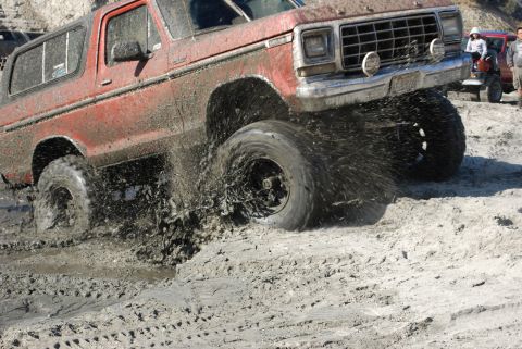 1996 Ford Bronco - A red Ford Bronco navigates a muddy off-road course, kicking up water and dirt.