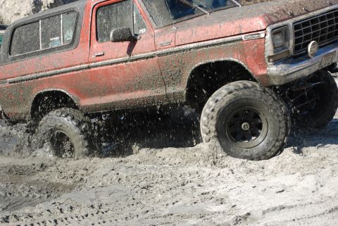 1996 Ford Bronco - This red SUV navigates a muddy terrain, with visible "SUPERMOTORS.NET" branding on the lower right.