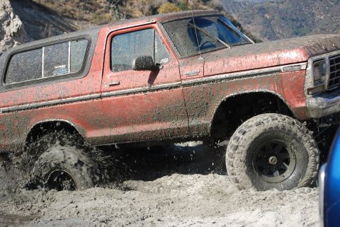 1996 Ford Bronco - Muddy red Ford Bronco navigating a challenging off-road terrain.