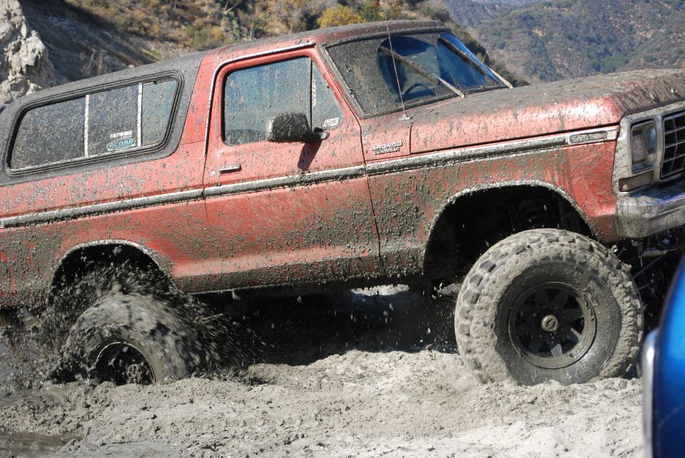 Red Ford Bronco with lifted suspension driving through thick mud, splashing water