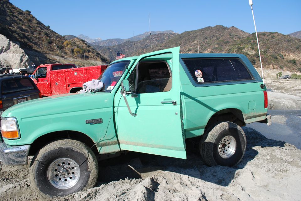 1996 Ford Bronco, driver's door and passenger door open, parked in sand with mountains in background. Red truck visible to the left
