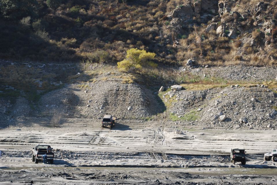 Three trucks on a dry, dusty landscape with hills in the background. One truck is in the foreground, another in the mid-ground, and a third further back