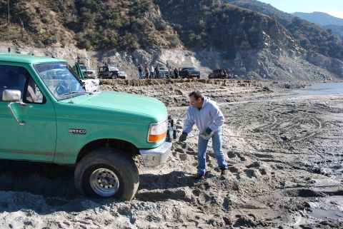 1996 Ford Bronco - Man attaches tow strap to green 1996 Ford Bronco in a dry lake bed with other vehicles present.