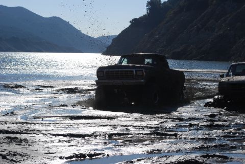 1996 Ford Bronco - Two trucks navigate a muddy terrain with a lake and hills in the background, kicking up mud and water.