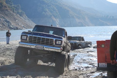 1996 Ford Bronco - Two trucks are stuck in the mud by the water's edge, with a person standing on the shore.