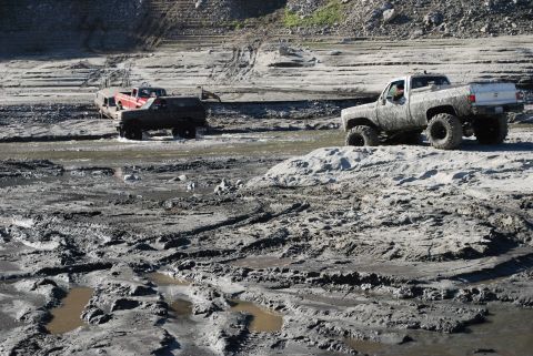 1996 Ford Bronco - Two trucks navigate a treacherous, muddy terrain in what appears to be a dry riverbed.