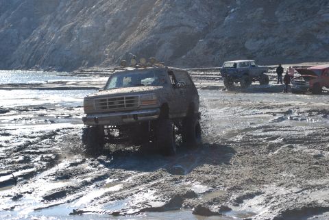 1996 Ford Bronco - An off-road SUV navigates a challenging muddy terrain, kicking up water and mud.