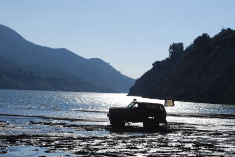 1996 Ford Bronco - A lone SUV sits on a muddy bank next to a shimmering lake, with dark mountains rising in the distance.