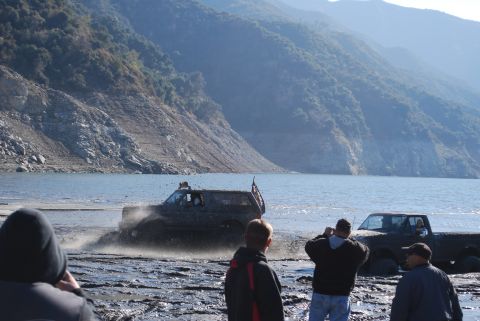 1996 Ford Bronco - Two off-road vehicles are seen in a muddy area near a large body of water, with spectators observing.