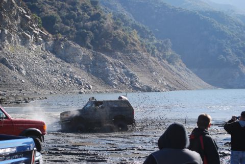 1996 Ford Bronco - Several vehicles are navigating a muddy terrain near a body of water, with spectators watching.