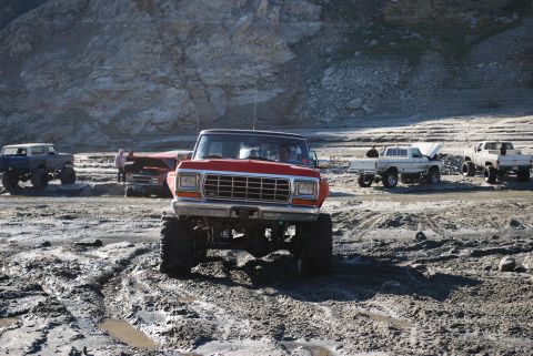 1996 Ford Bronco - Several off-road trucks are gathered in a muddy, rocky area, with one red truck prominently in the foreground.