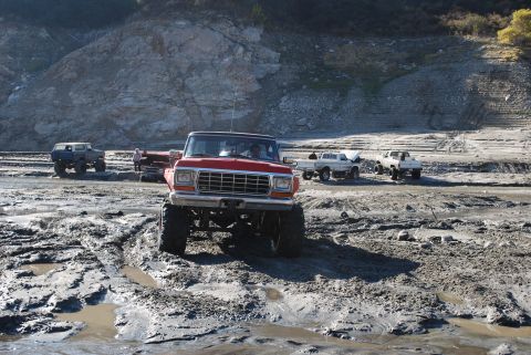1996 Ford Bronco - Several off-road vehicles navigate a muddy terrain under a rocky hillside.