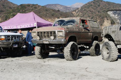 1996 Ford Bronco - Two heavily modified, mud-caked SUVs are parked outdoors under a purple tent.