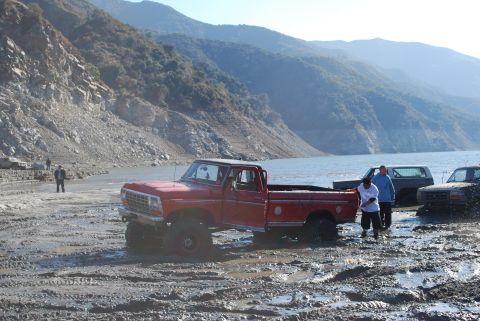 1996 Ford Bronco - Several vehicles are navigating a muddy, off-road area near a large body of water and hills.