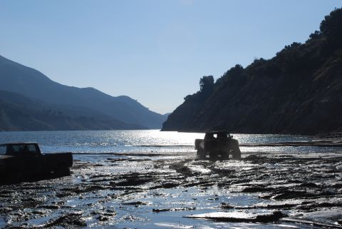 1996 Ford Bronco - Two trucks navigate a muddy, wet terrain with a lake and mountains under a clear sky.