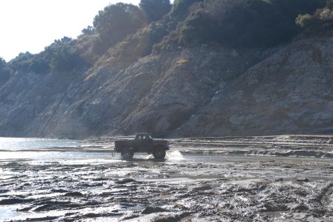 1996 Ford Bronco - A dark pickup truck navigates through a muddy, watery terrain with a steep, tree-covered hillside under a bright sky.